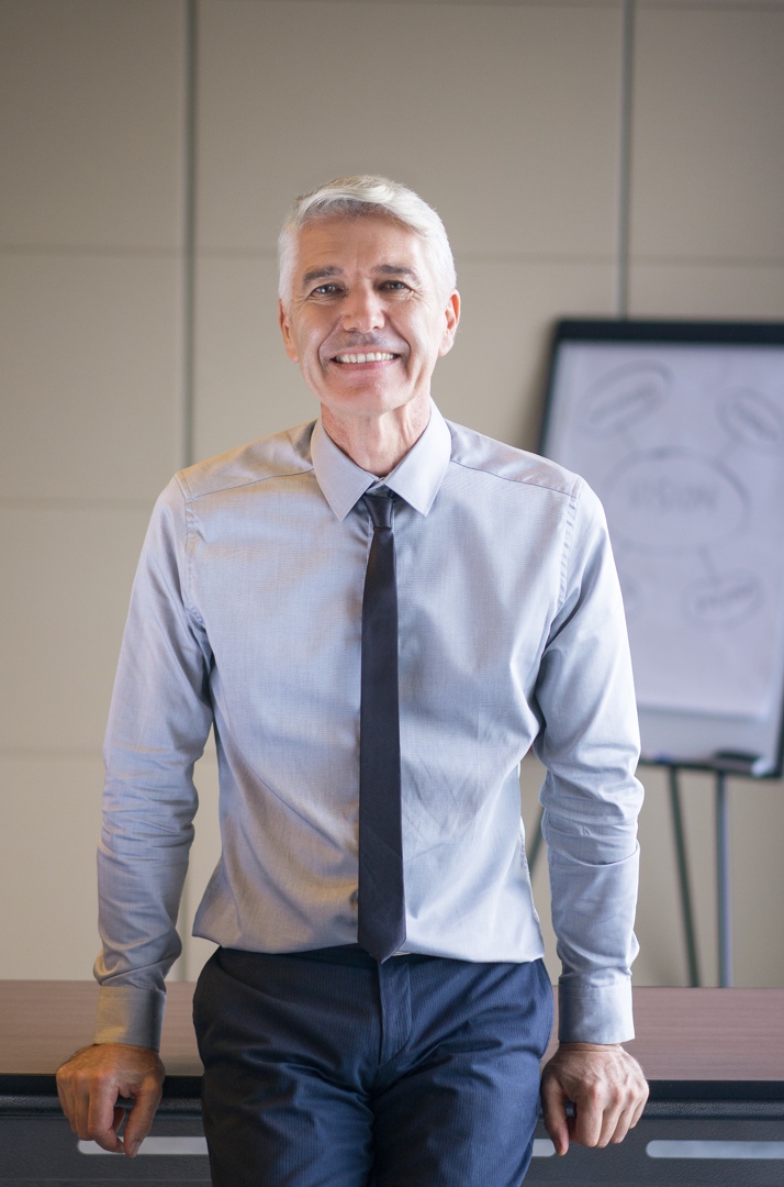 Closeup portrait of smiling senior businessman looking at camera and leaning on desk with blurry flipchart in background